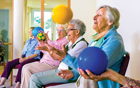 A group of people are in a room, some of them are sitting on chairs, and others are standing. A man is kneeling down next to a woman who is sitting on the floor. The group appears to be engaged in an exercise or physical activity. AI generated content